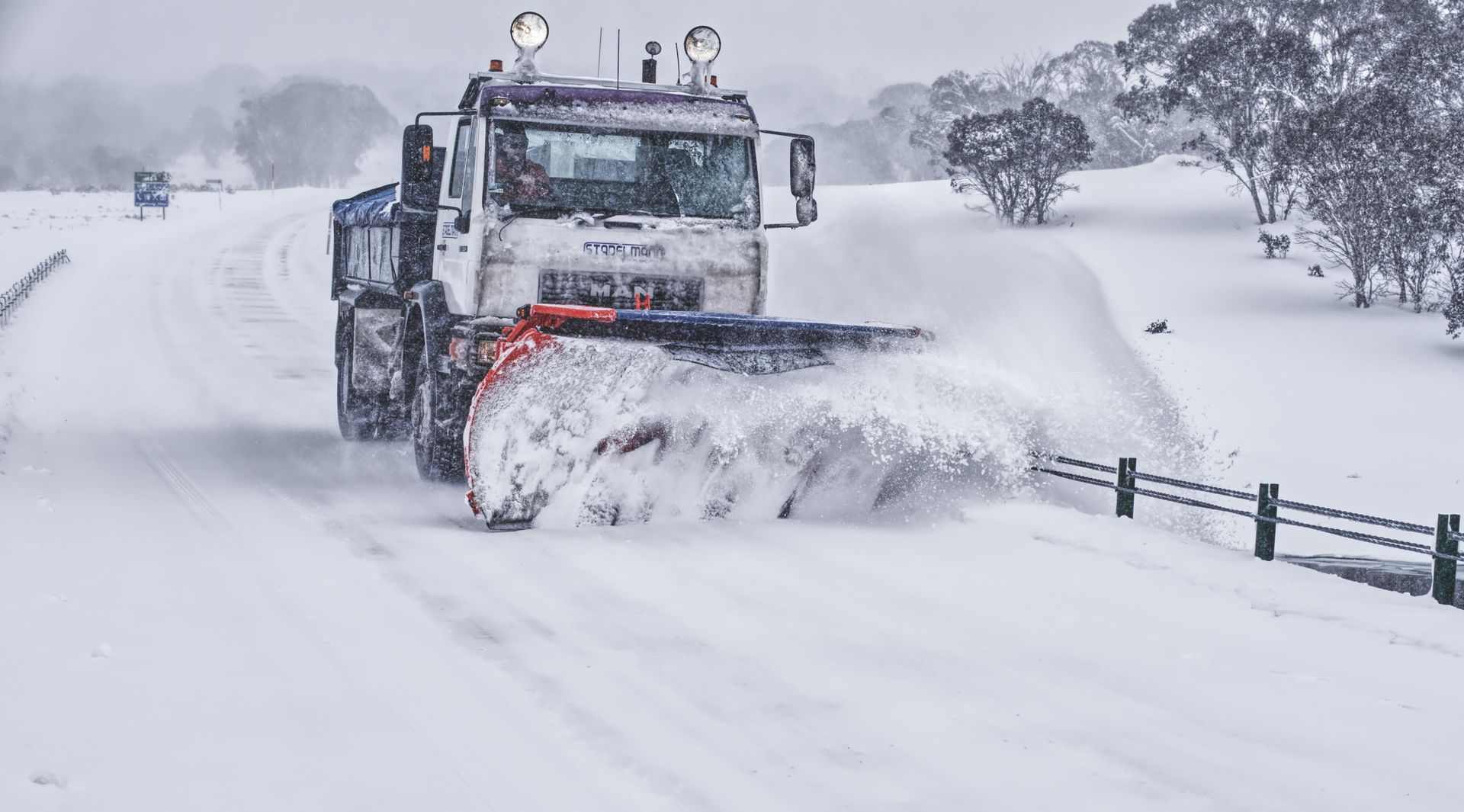 Snowplow clearing a snowy road with trees in the background.