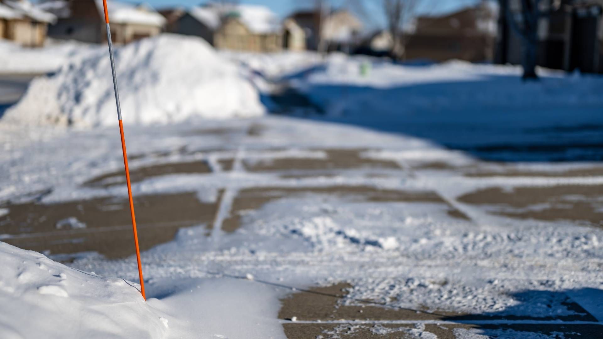 Snow-covered driveway in a residential area, with a pile of snow and an orange marker.