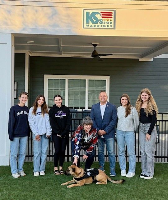 A group of people posing with a dog in front of a building that says ks