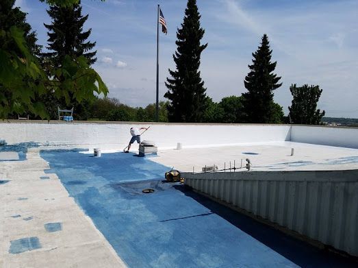 Person painting a flat roof blue and white, with flag pole and trees in the background.