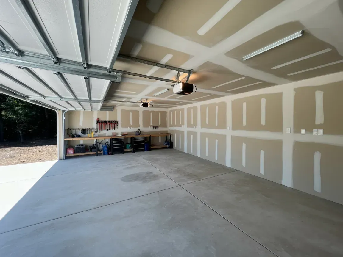 Open, empty garage with a concrete floor, drywall walls, and open garage door.