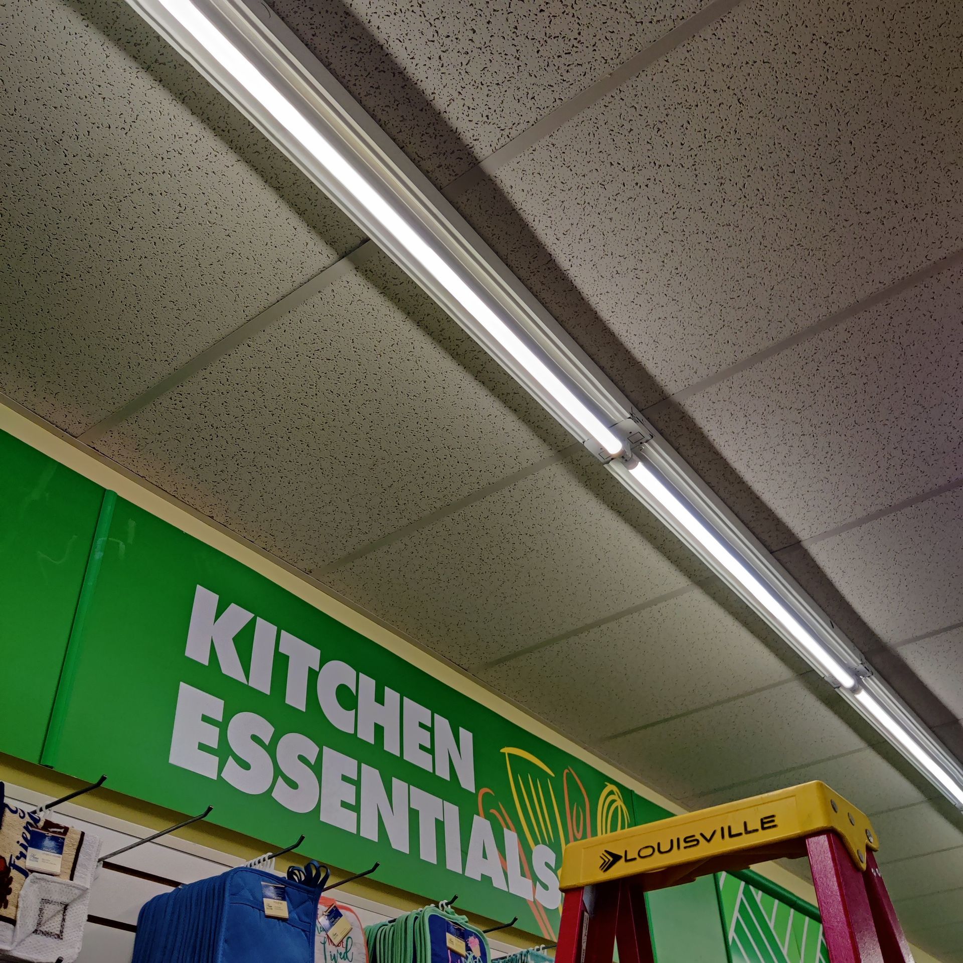 Store aisle with green “Kitchen Essentials” signage and fluorescent ceiling lights, plus a yellow display sign.