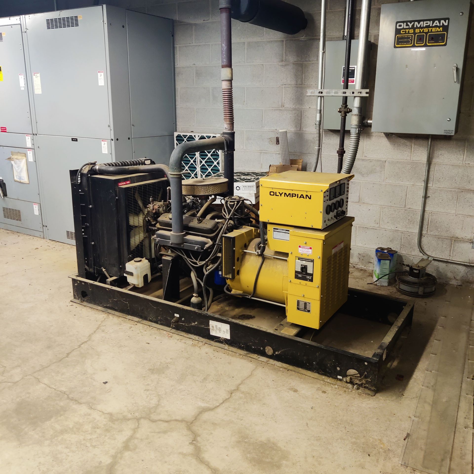 Industrial generator set on a black skid in a utility room, with gray electrical cabinets and concrete walls.