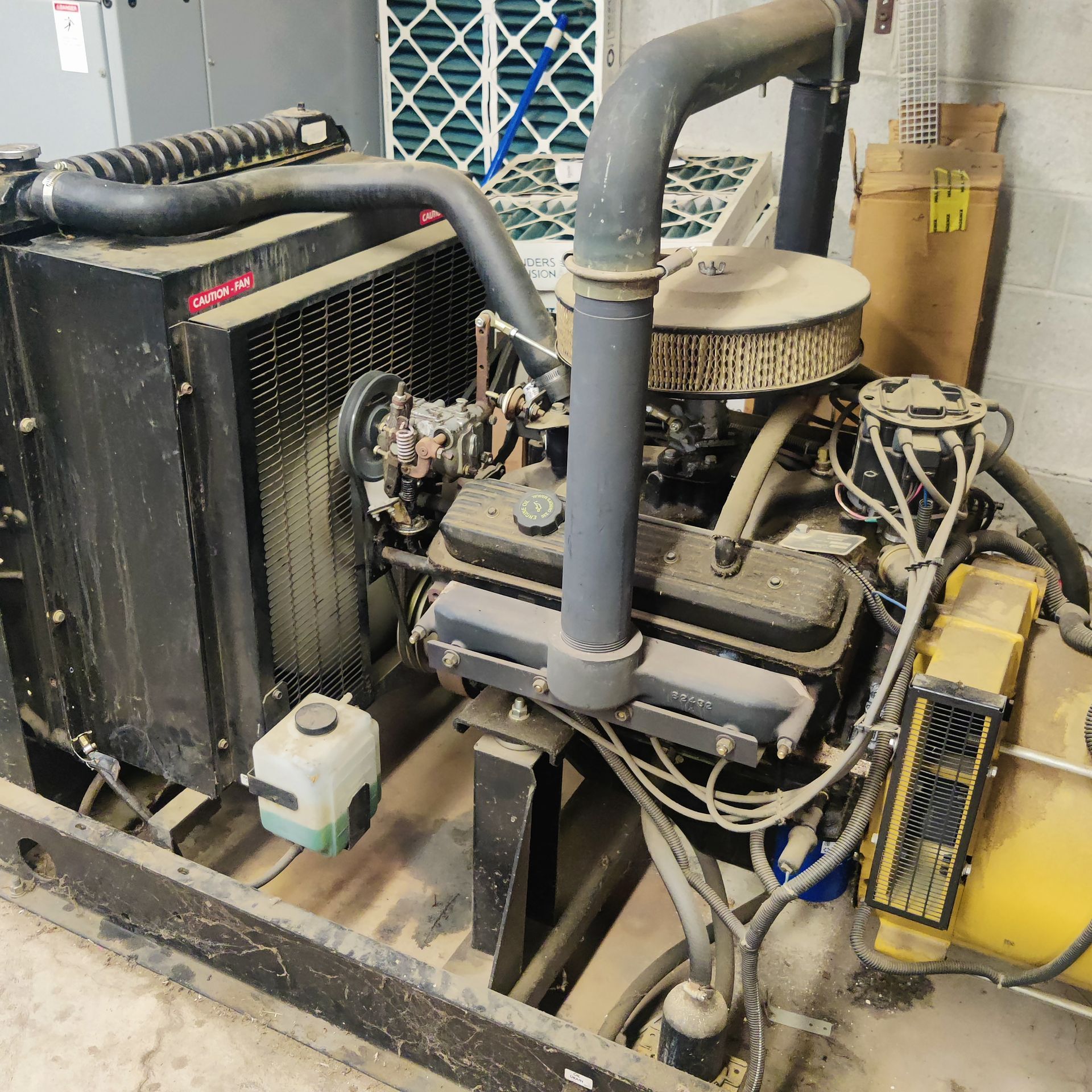 Industrial engine/generator unit with black radiator, pipes, and cables in a dusty mechanical room