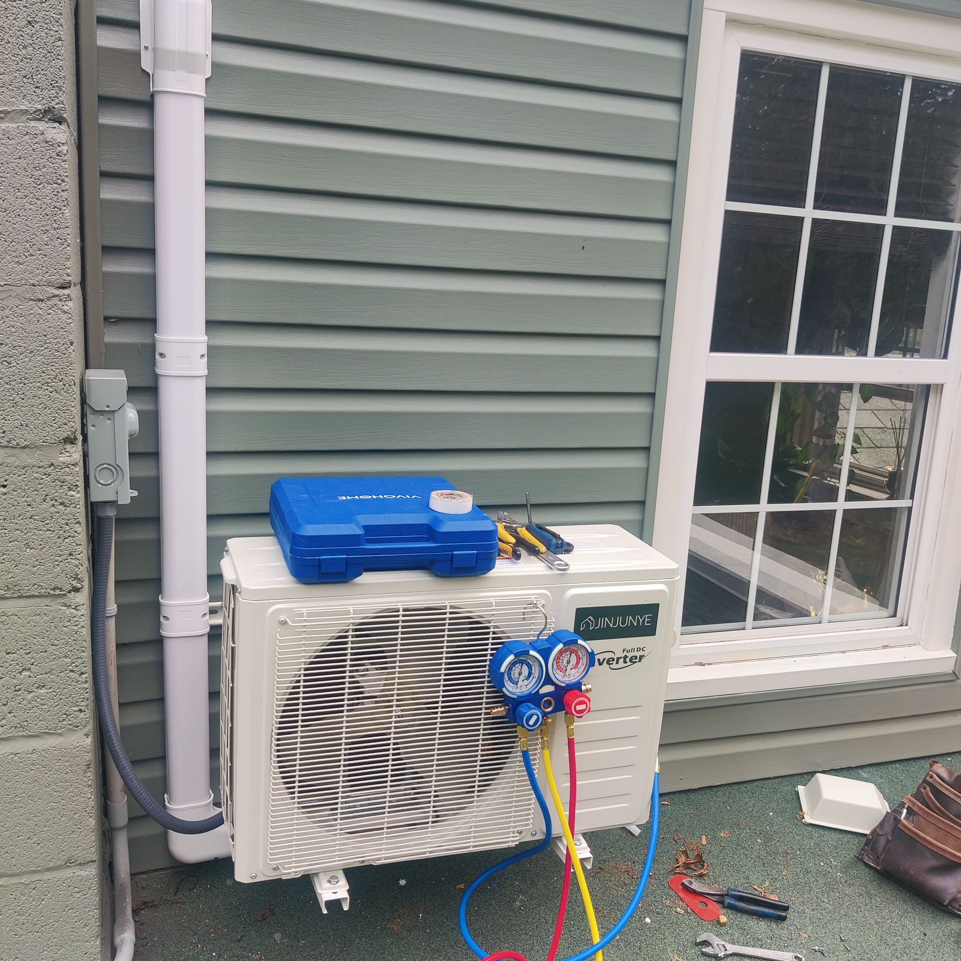 Outdoor HVAC unit beside house siding, with blue manifold gauges and tools attached to the front.