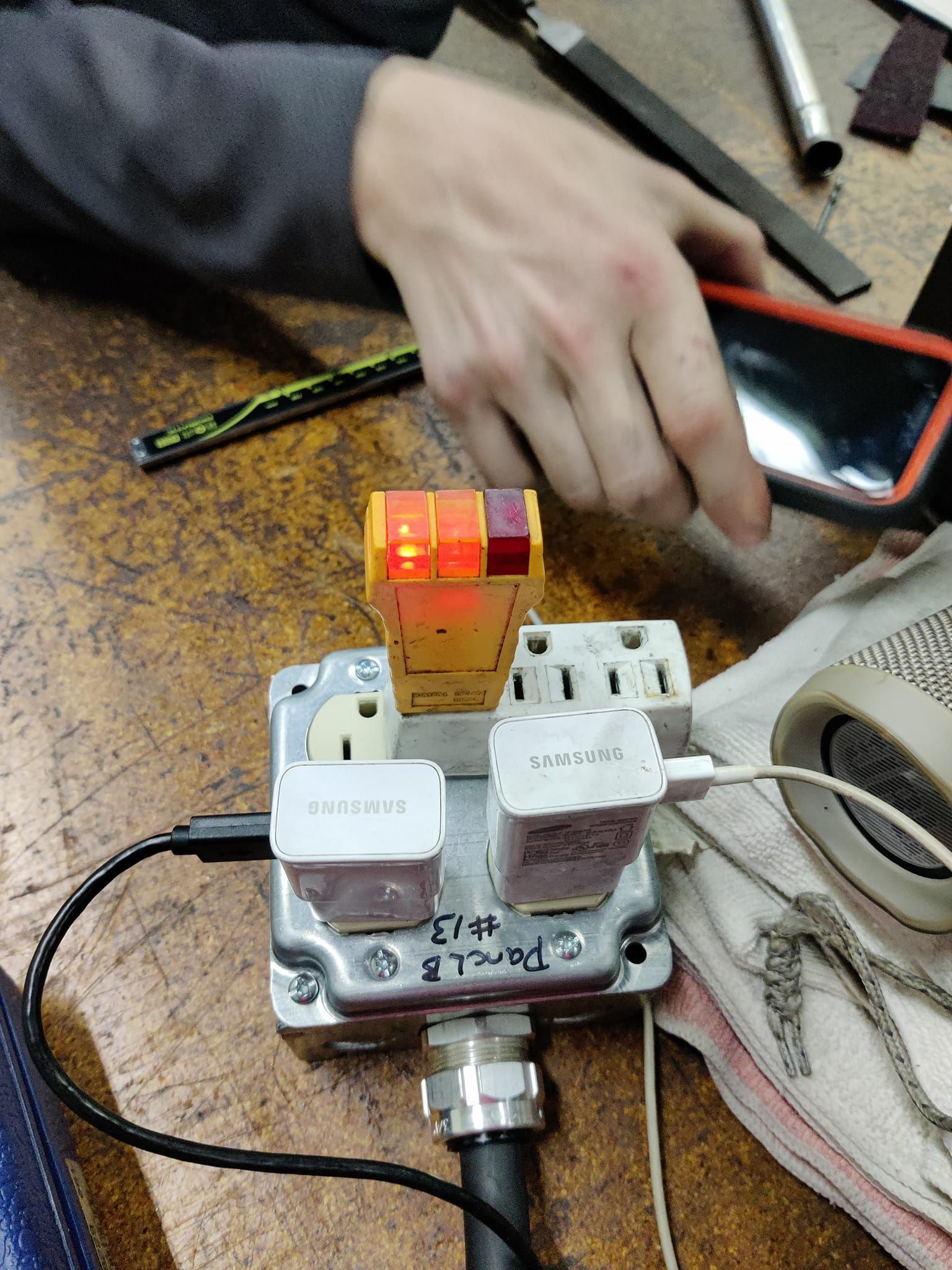 Hand using a small electronic device with a glowing red light on a workbench, with cables attached