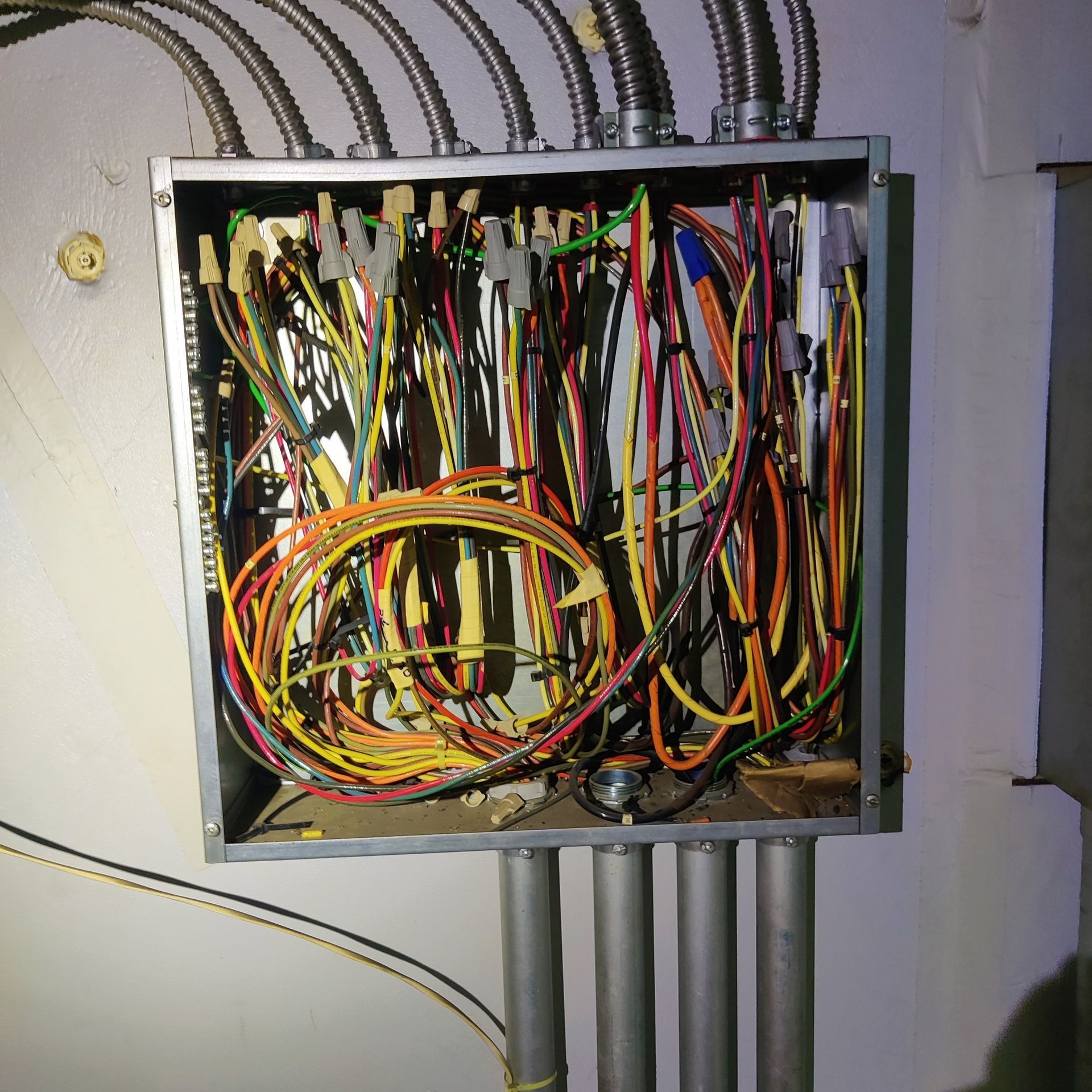 Open electrical junction box with tangled multicolored wires and conduit pipes.