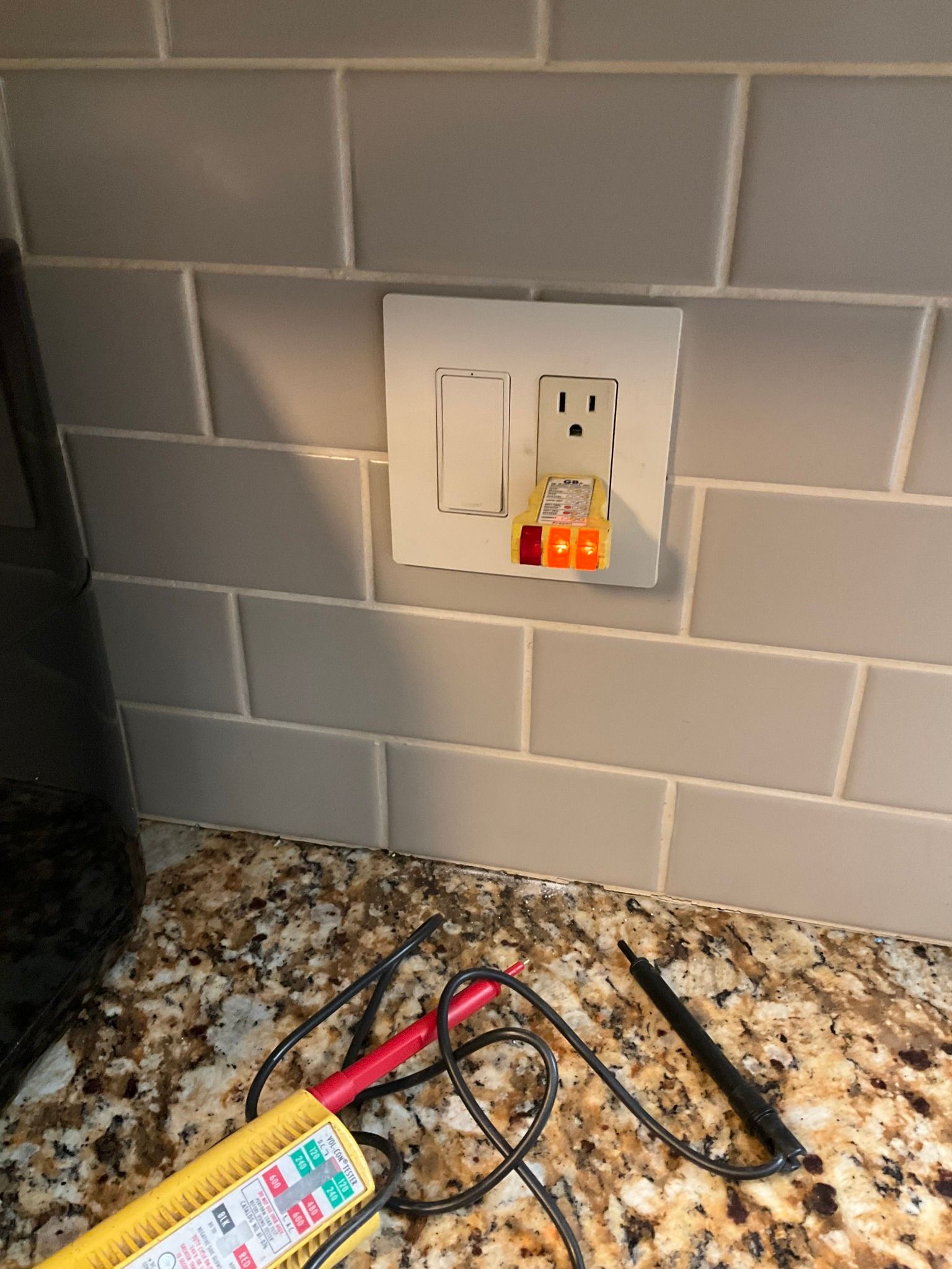 Kitchen outlet with a plug-in outlet tester on a granite countertop, next to a black cord and a multimeter probe.