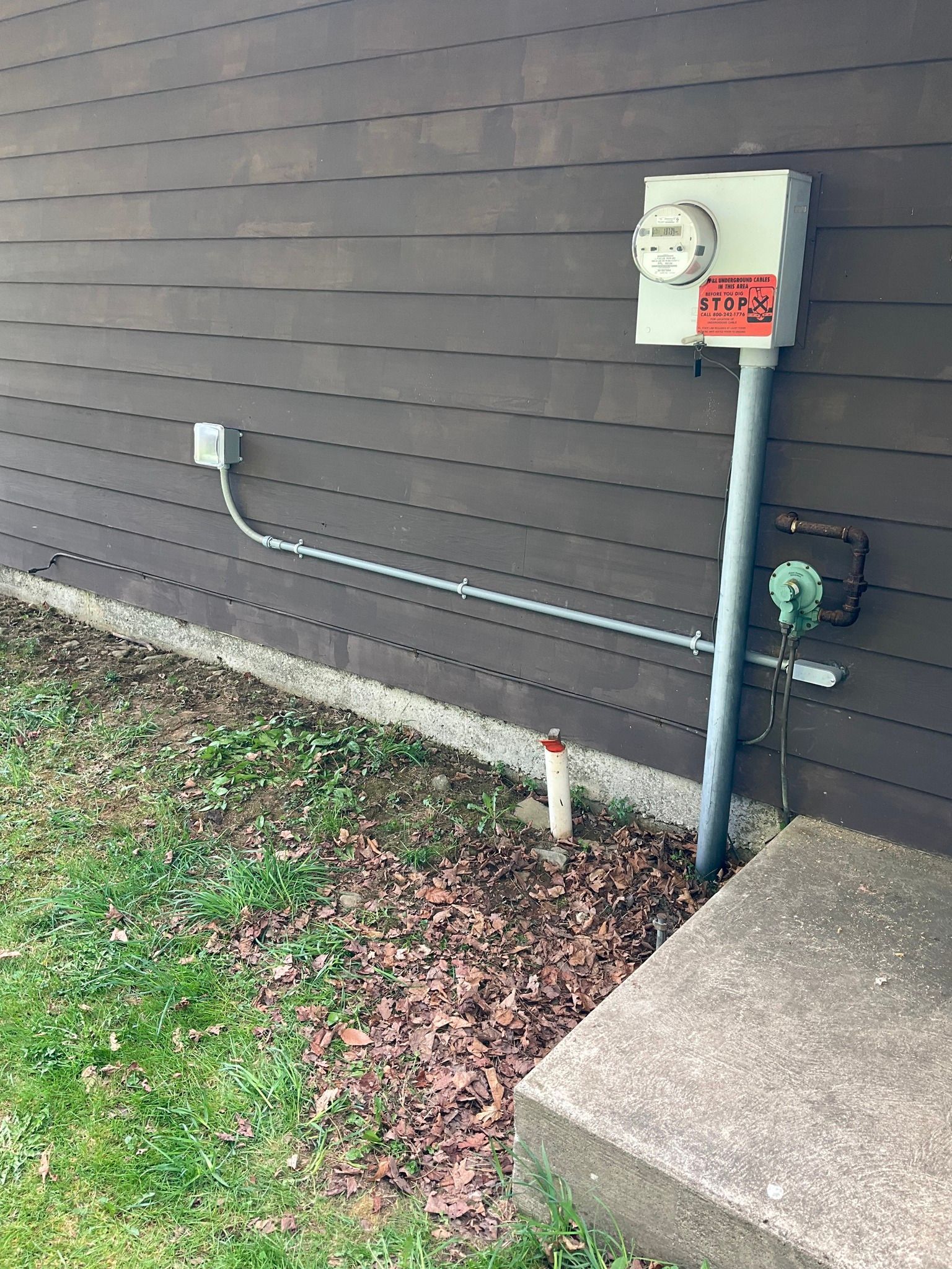 Utility meter and electrical conduit on a brown building wall beside a concrete step and patch of grass and leaves