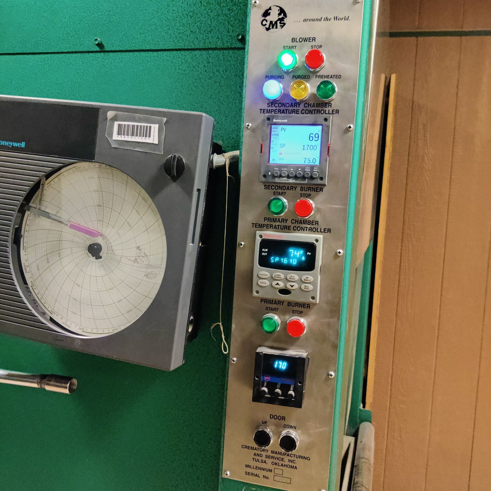 Industrial control panel with lit buttons and digital displays beside a circular chart recorder on a green wall