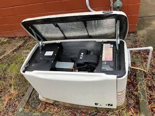 Open utility generator outside a brick building with lid raised, revealing internal components and controls.