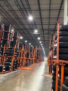 Warehouse aisle with tall orange pallet racks and stacked black tires under bright ceiling lights