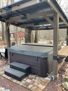 Covered outdoor hot tub with steps on a stone patio in a wooded setting