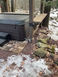 Snow-dusted patio beside a hot tub and wooden deck with stone edging in a wooded yard