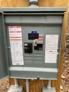 Gray electrical service panel mounted on plywood, with warning labels and meter in a utility area