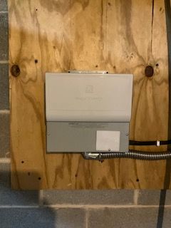 White electrical panel mounted on plywood wall above cinder blocks, with conduit and cable attached.