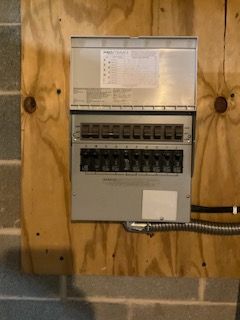 Electrical breaker panel mounted on plywood wall, with exposed wiring and multiple switches.