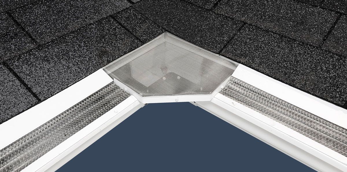 A close-up of a white corner guard with a metallic plate installed on the corner of a dark, shingled roof edge.