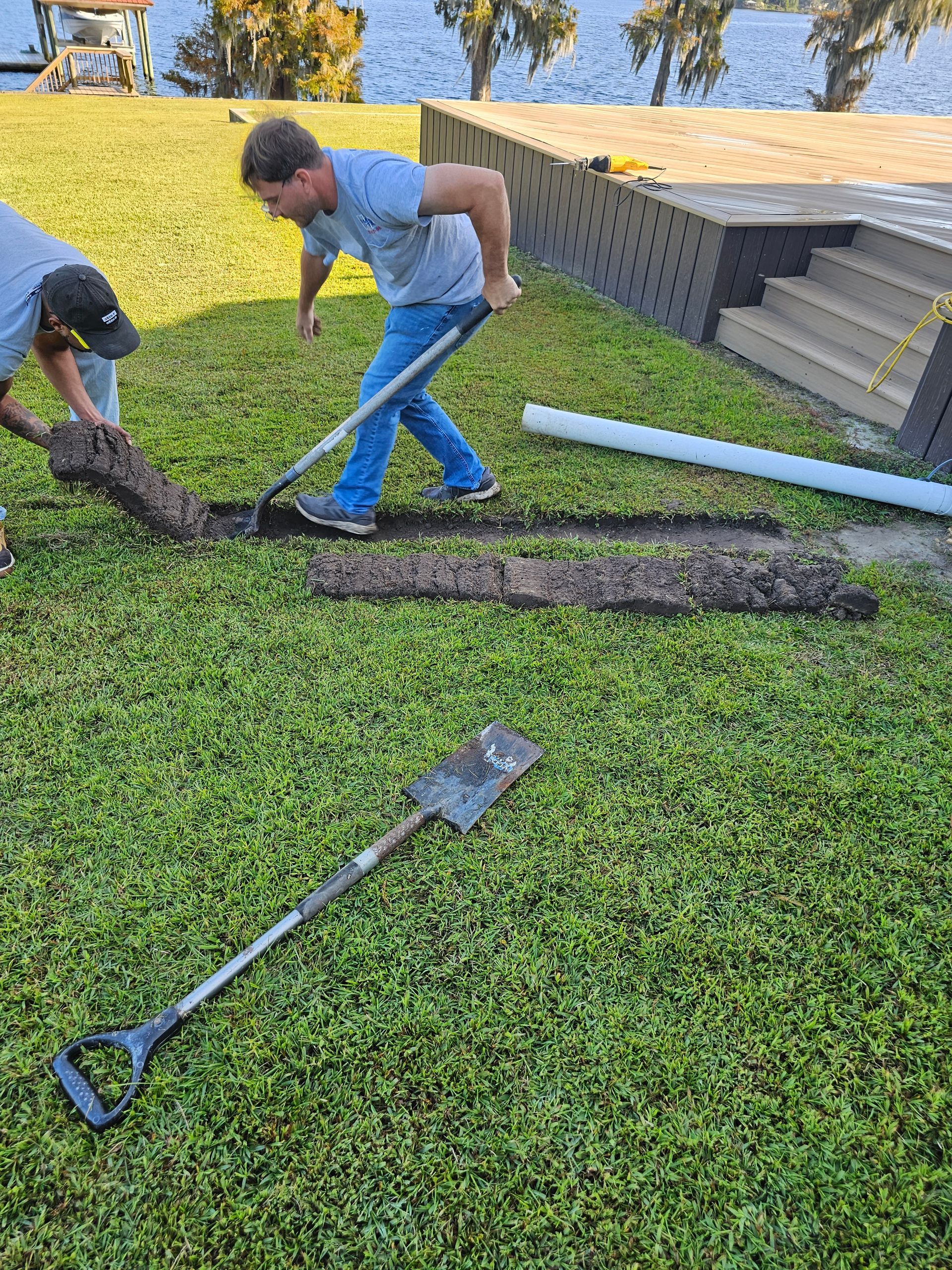Two men digging a trench in green grass next to a lake; one uses a shovel, the other, a rake.