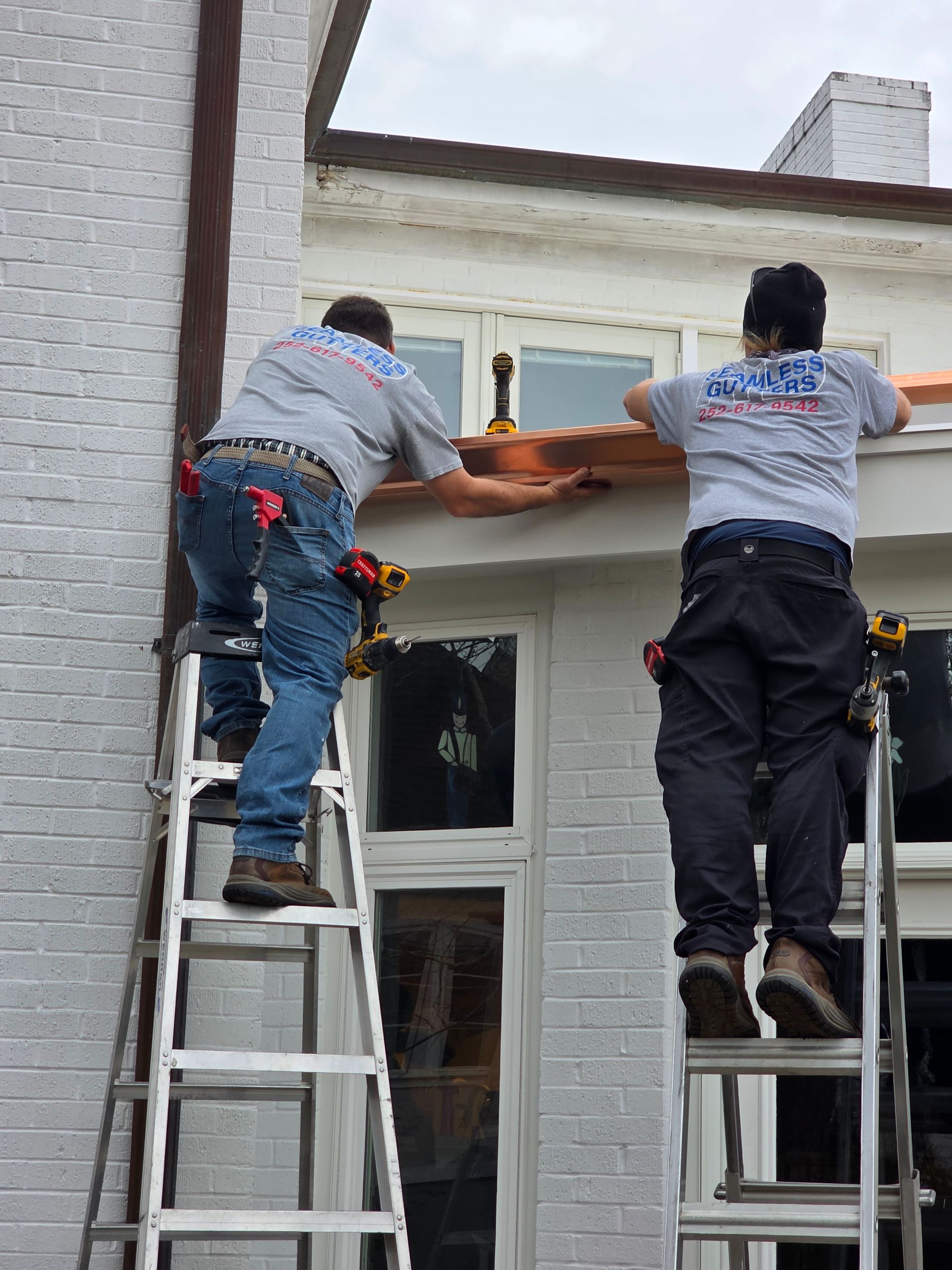 Two workers in grey uniforms stand on separate ladders while installing copper flashing along the roofline of a home.