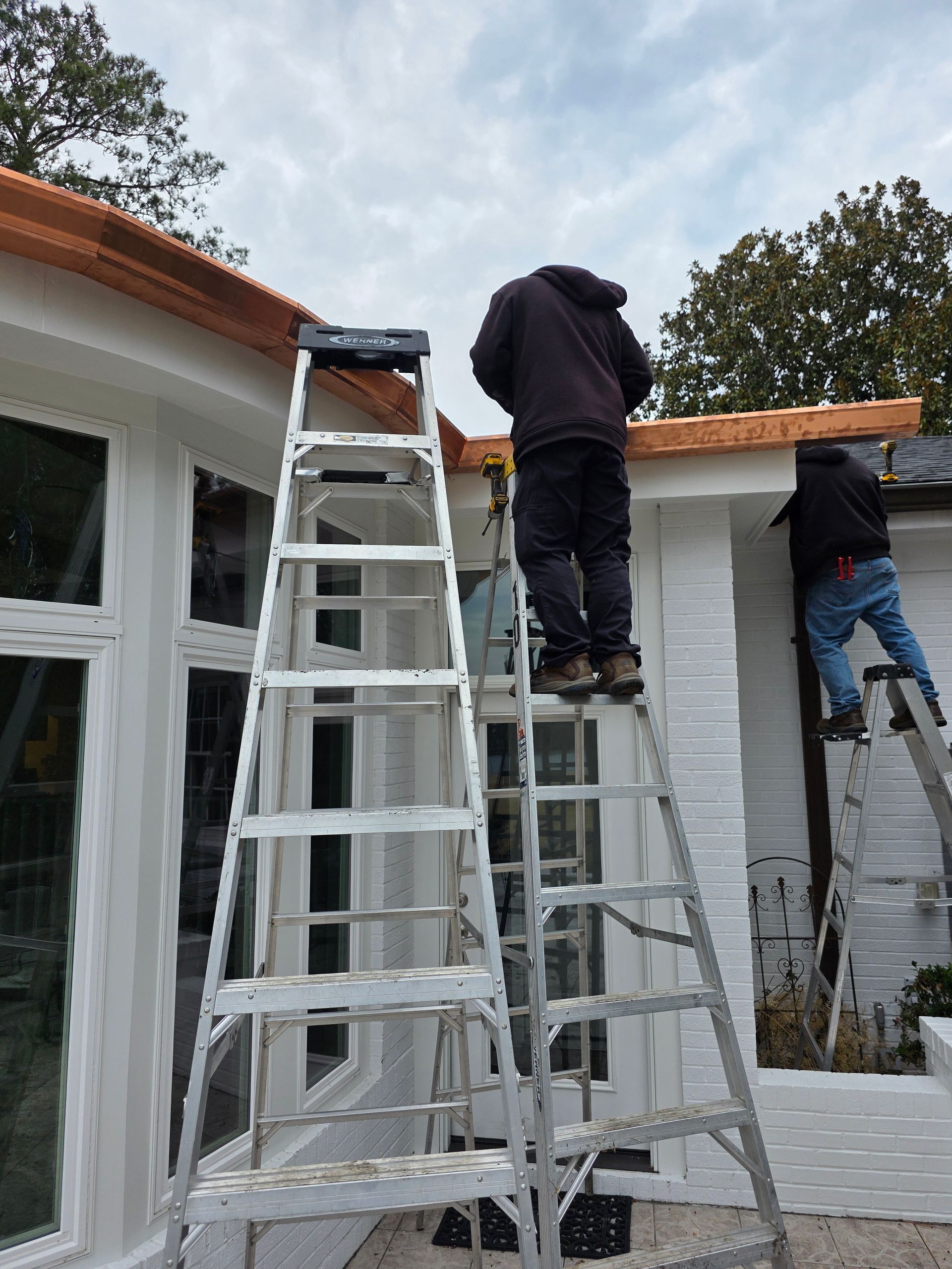Two workers on ladders install wooden trim along the roofline of a white-painted brick home.