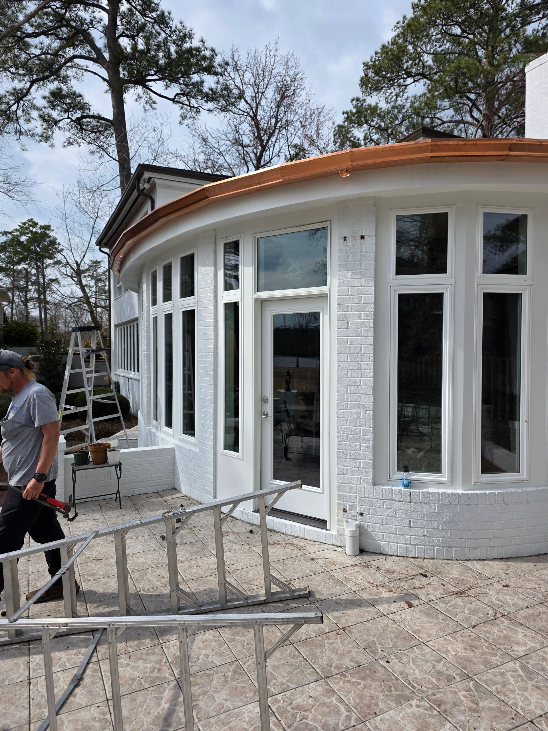 A worker stands near a ladder by a freshly painted white, curved building with large windows on a stone patio.