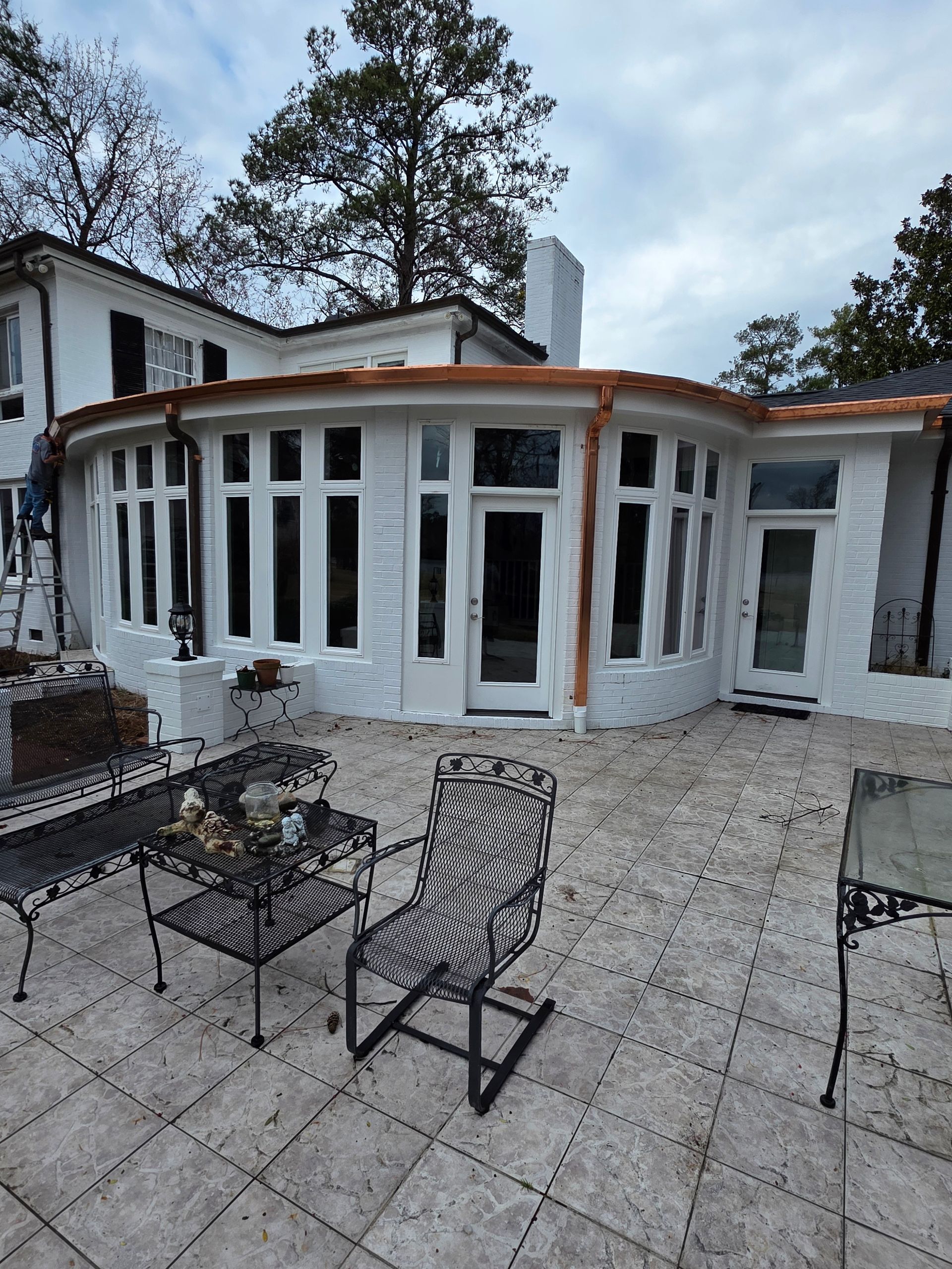 A white sunroom with a curved wall and copper trim on a patio with metal furniture and a person on a ladder.