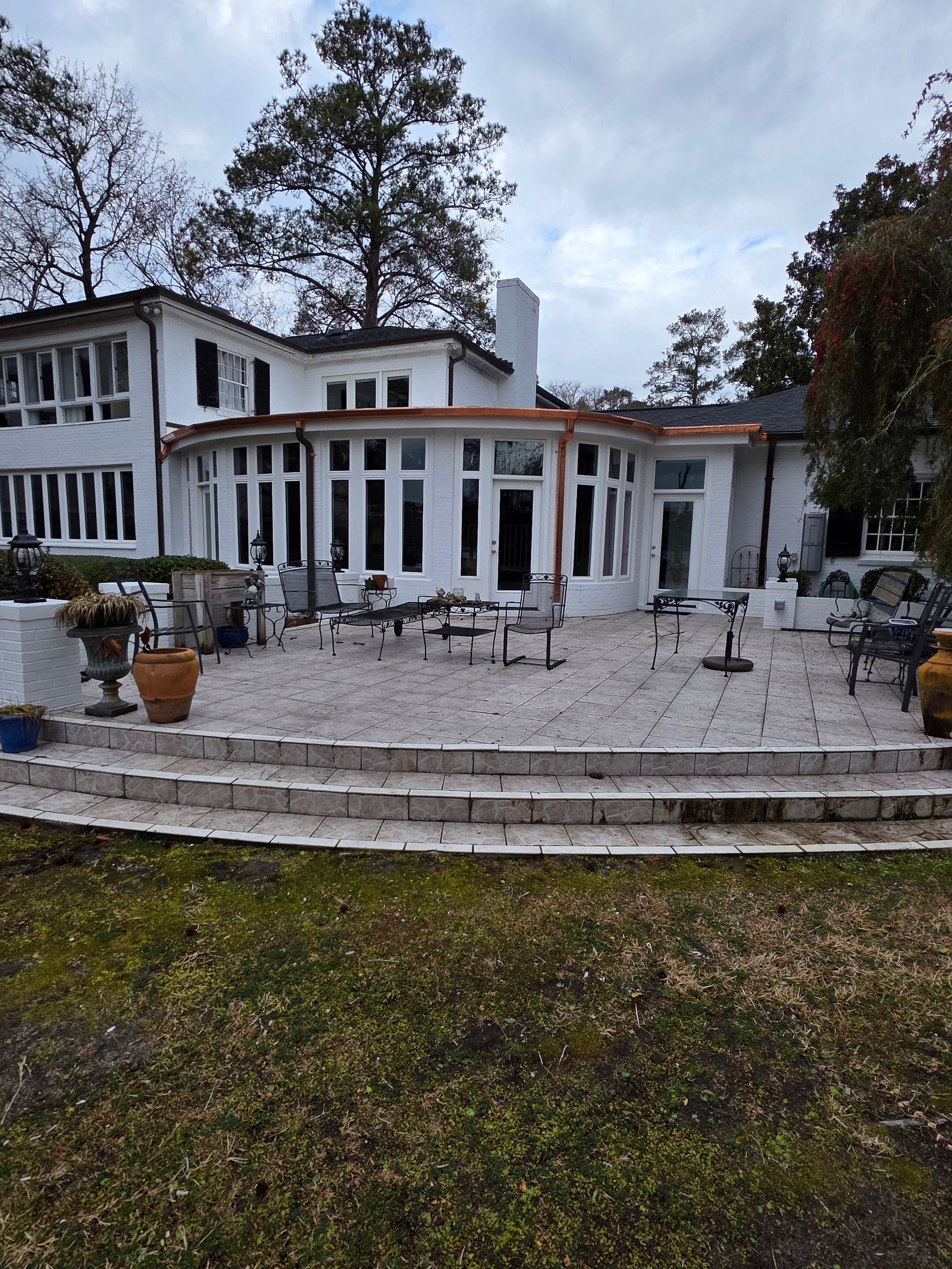 A white multi-story house with a curved sunroom, brick patio, and tiered steps leading to a lawn under a cloudy sky.