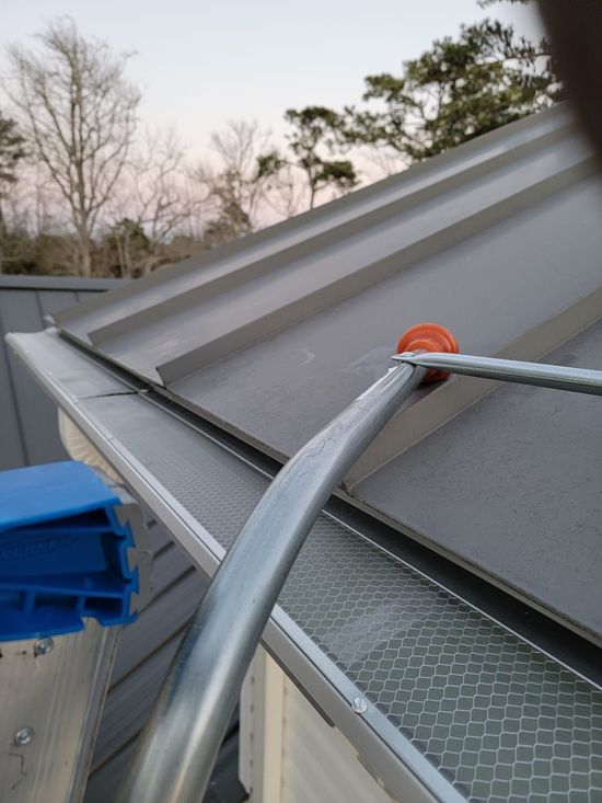 Gutter with leaf guard on a gray metal roof. A ladder and a hand are visible.