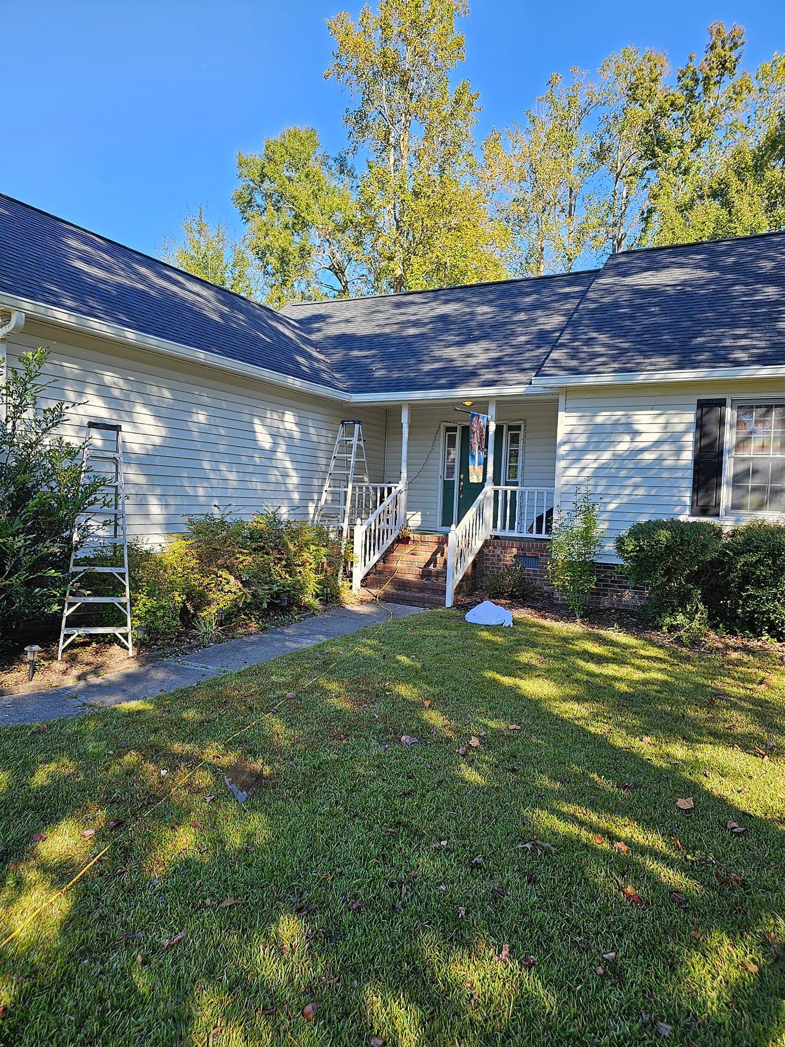 White house with dark roof, green bushes, and ladder on the lawn. Clear, sunny day.