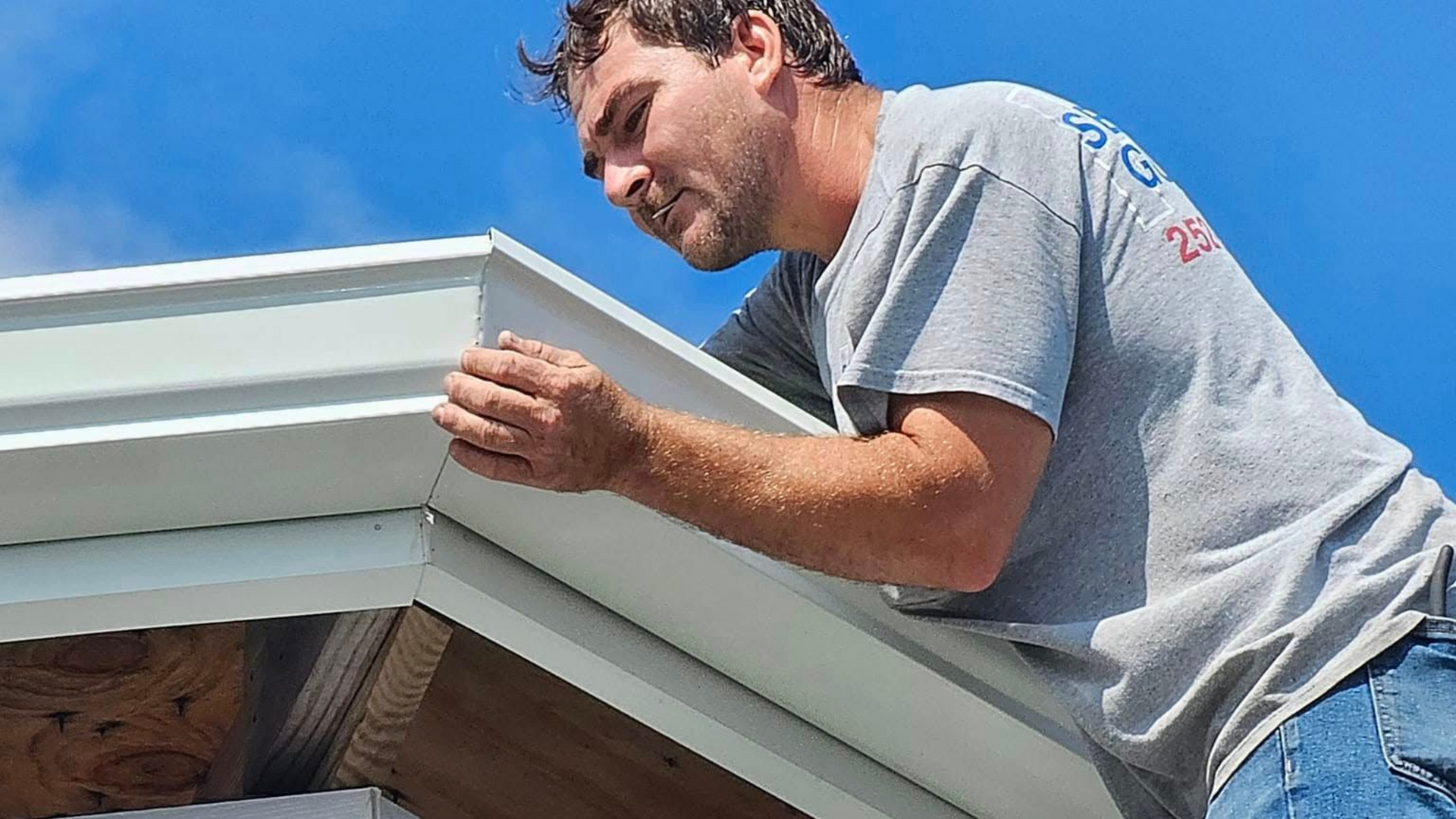 A worker installs white metal flashing on the edge of a roof against a clear blue sky.