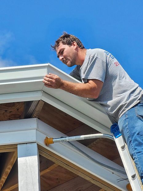 Man on ladder installing white trim on a roof under a blue sky.