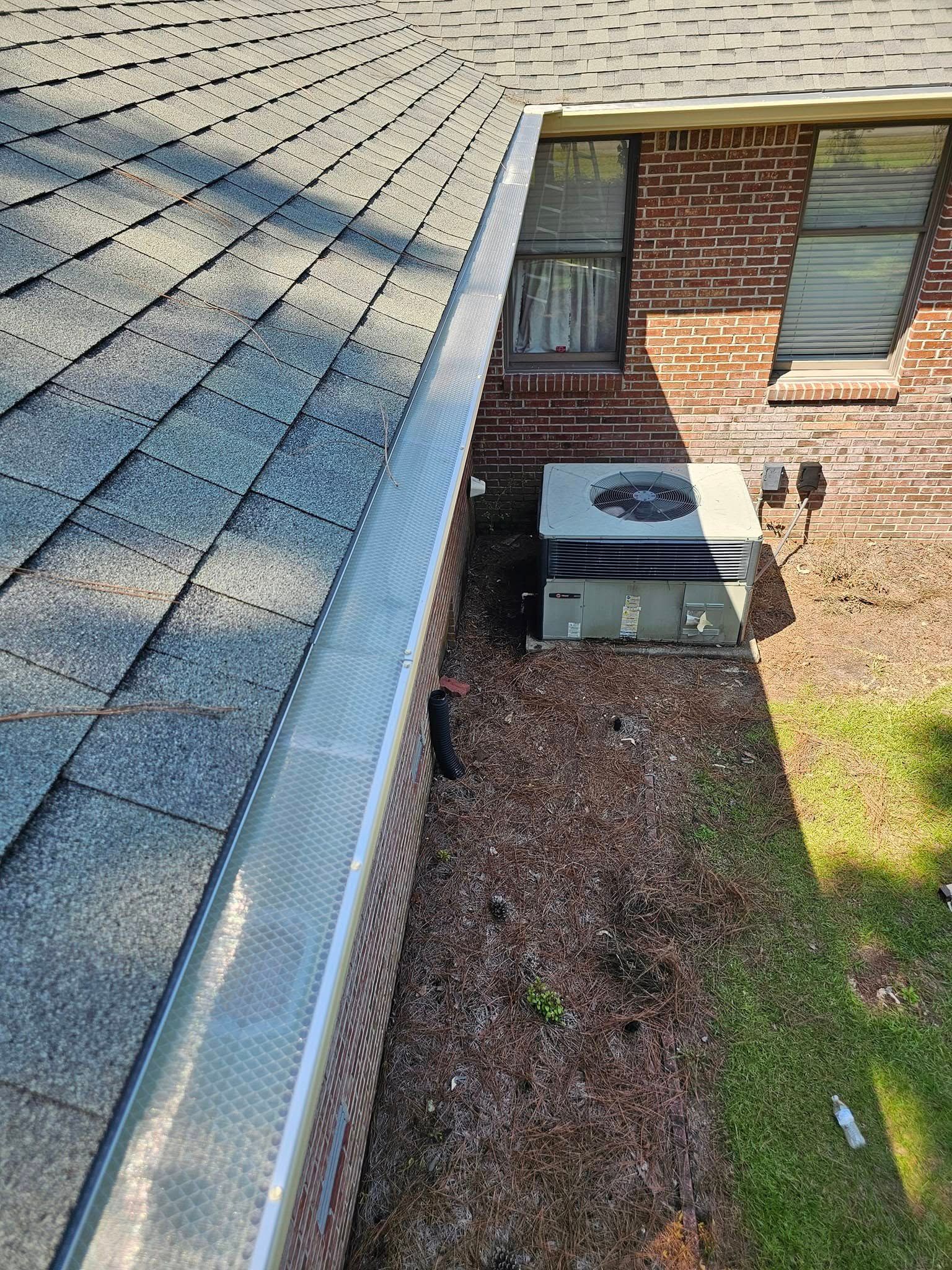 A roof with gray shingles and a silver gutter next to a brick wall, air conditioning unit and grass.