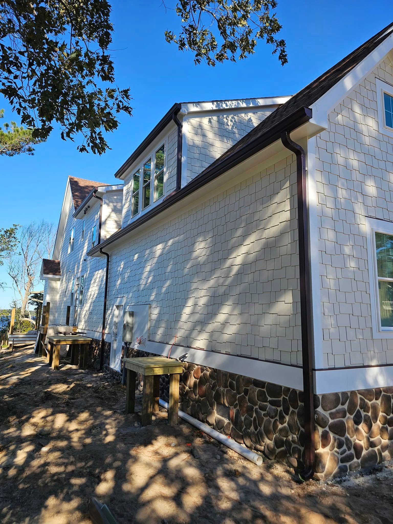 Two-story house under construction, white siding, stone base, brown trim, clear blue sky.