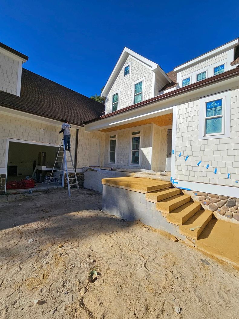 A person on a ladder works on the white exterior of a new house with unpaved ground, stairs, and a clear blue sky.