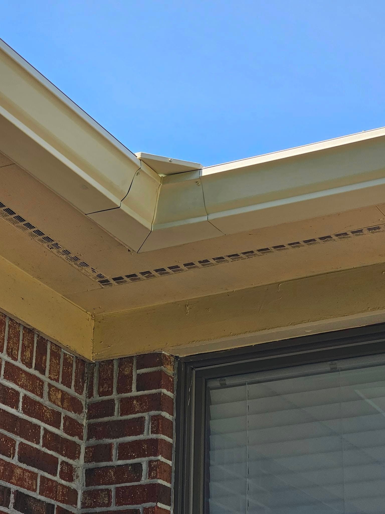 Corner of a brick building with beige eaves and a window, against a blue sky.