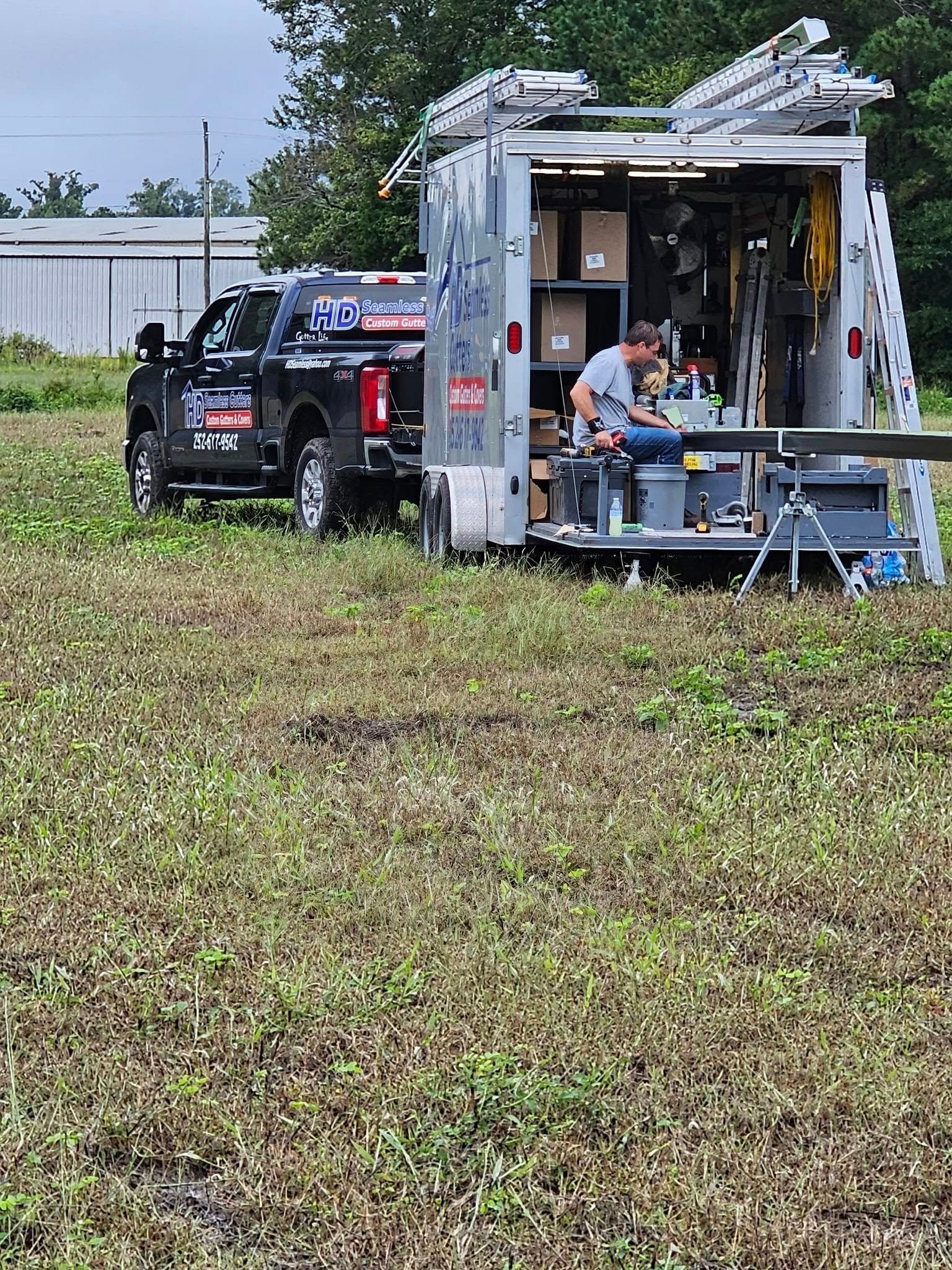 A truck pulling a trailer, a man working on equipment outside. Located in a field.