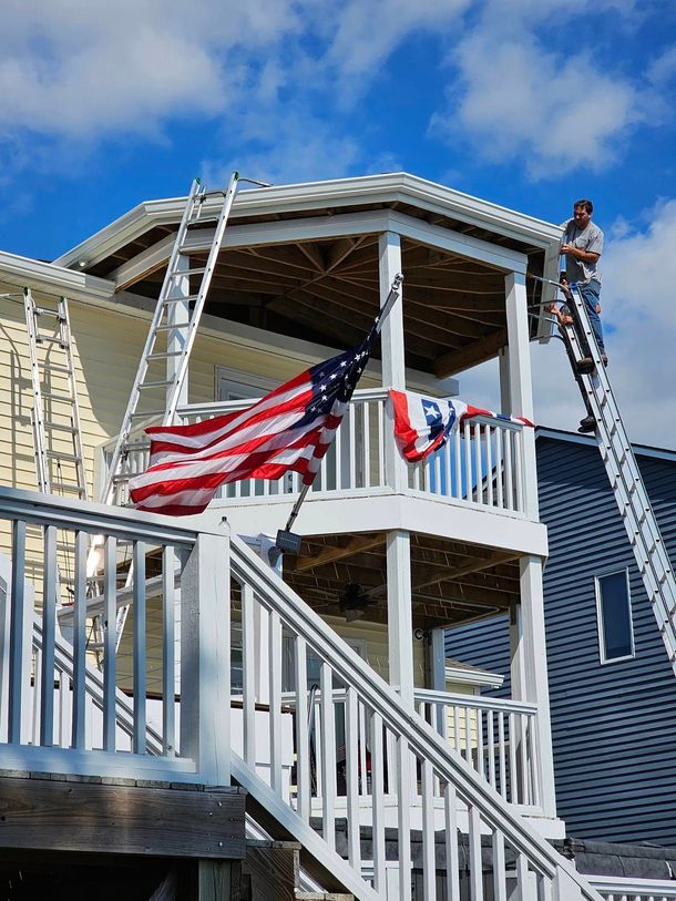 American flags hang from a white porch; a person on a ladder works on the roof.