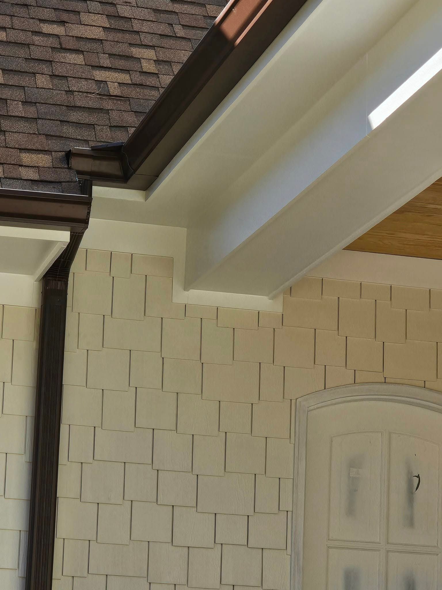 Close-up of a house exterior with a brown roof, tan siding, and white trim.