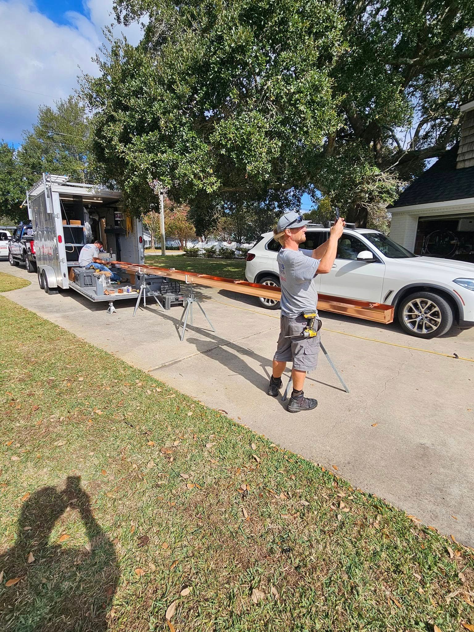 A person holding a pole filming next to a trailer, car, and tree on a sunny day.