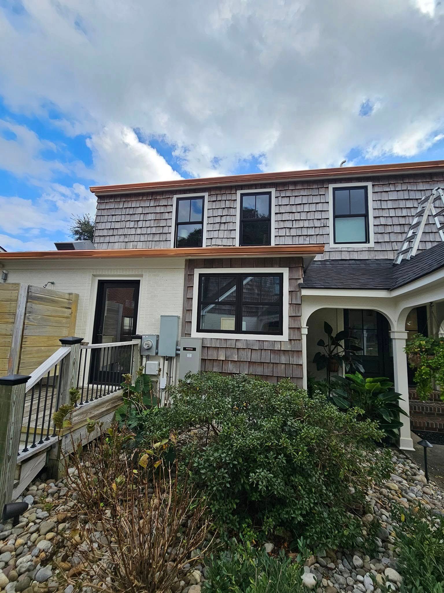 House exterior with gray shake siding, black-framed windows, and copper gutters. Green shrubs in foreground.