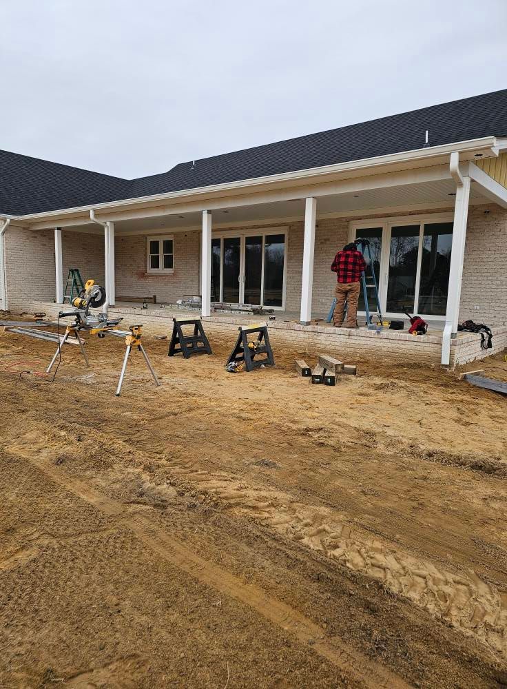 Construction site. Man working on a porch with sliding glass doors. Sawhorses, tools, and materials visible.