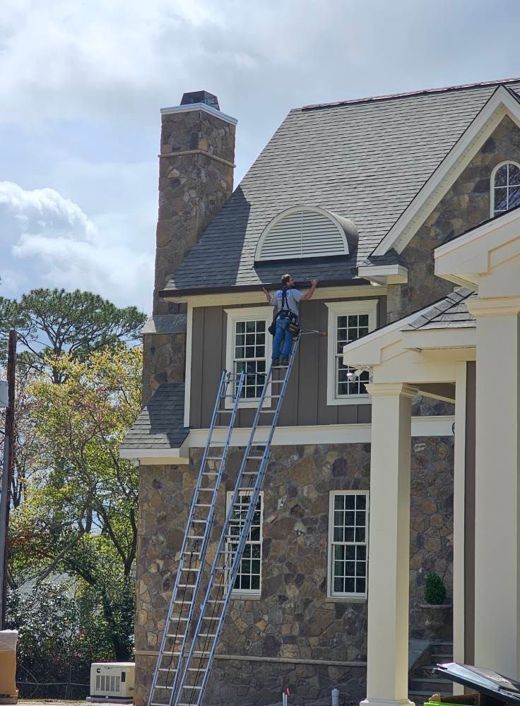 Man on a ladder working on a house's roof and siding. Stone facade, brown and gray tones.