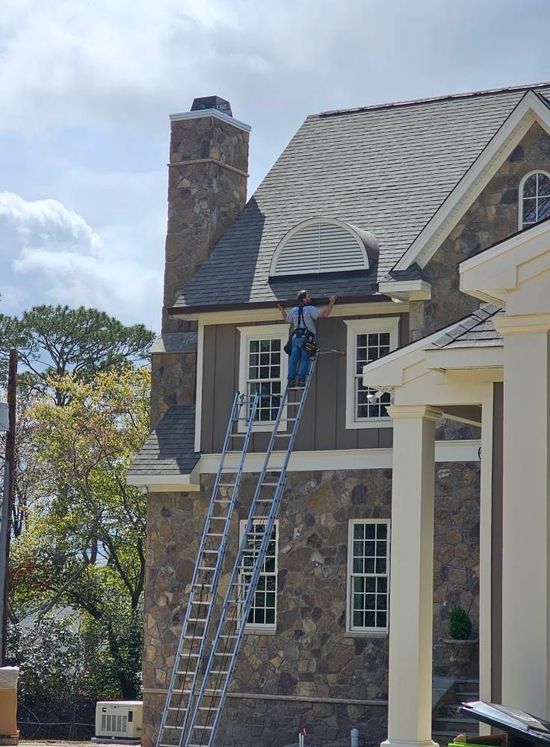Man on a tall ladder working on the side of a stone and wood house near the roof.
