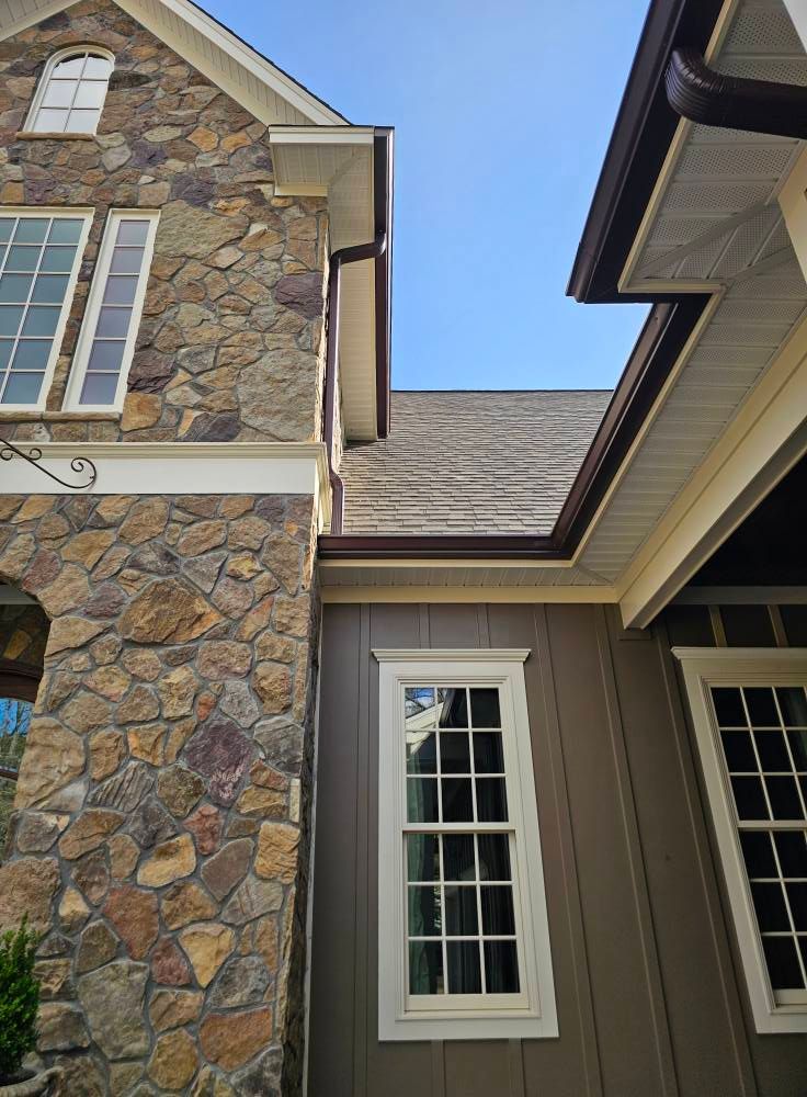 Exterior of a house with stone and gray siding, white trim, and brown gutters against a blue sky.