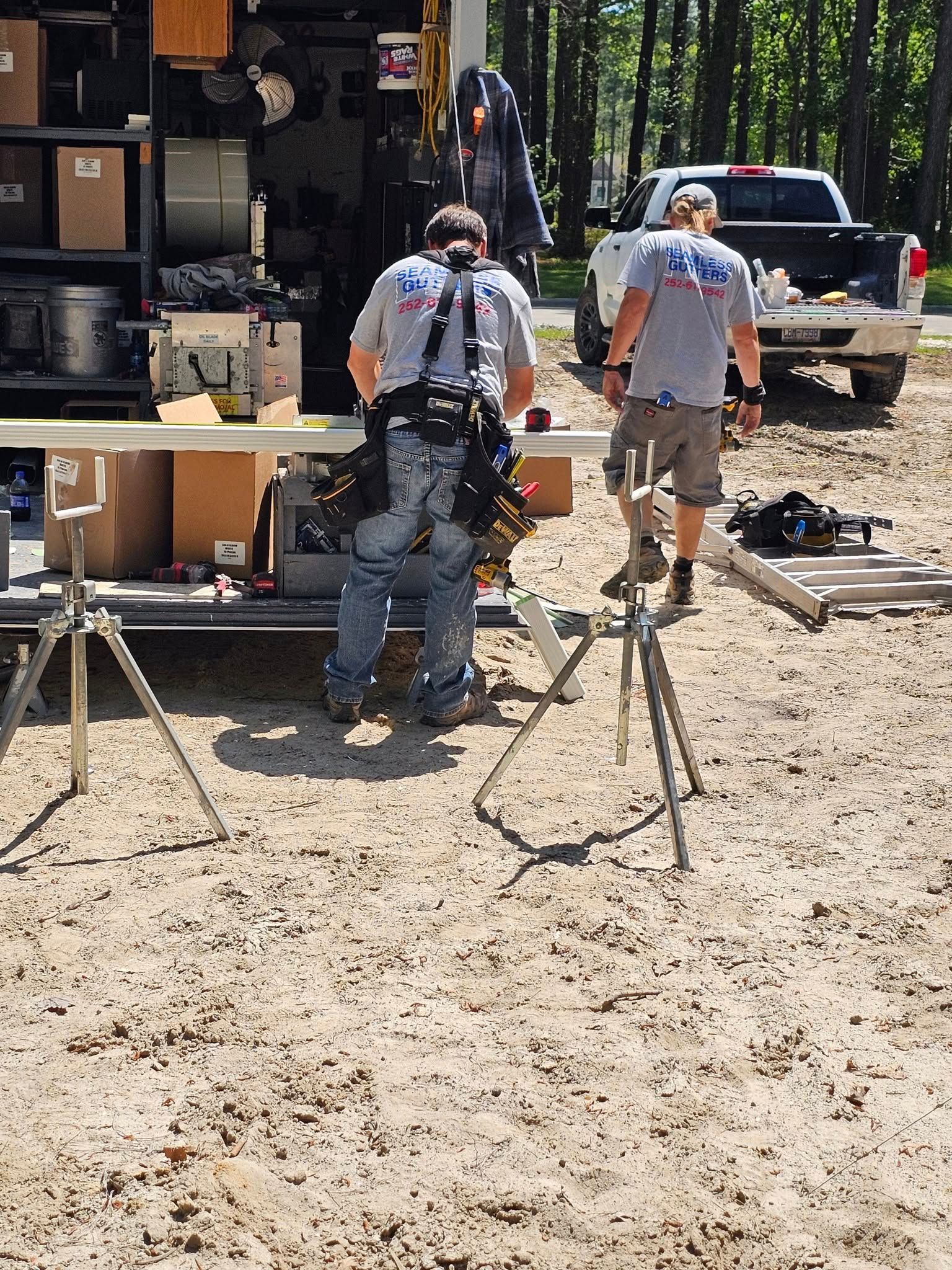 Two construction workers outside, one using a saw, near a truck with tools on a sandy site.