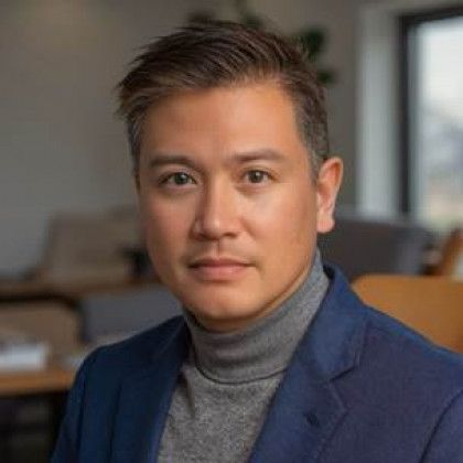 Man with short, dark hair, wearing a gray turtleneck and blue blazer, looking towards the viewer.