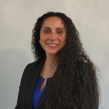 Woman with long, curly dark hair smiles, wearing a blue top and black blazer, against a white wall.