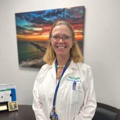 Woman in a white coat smiles, wearing glasses. She stands in an office with a painting of a coastal scene.