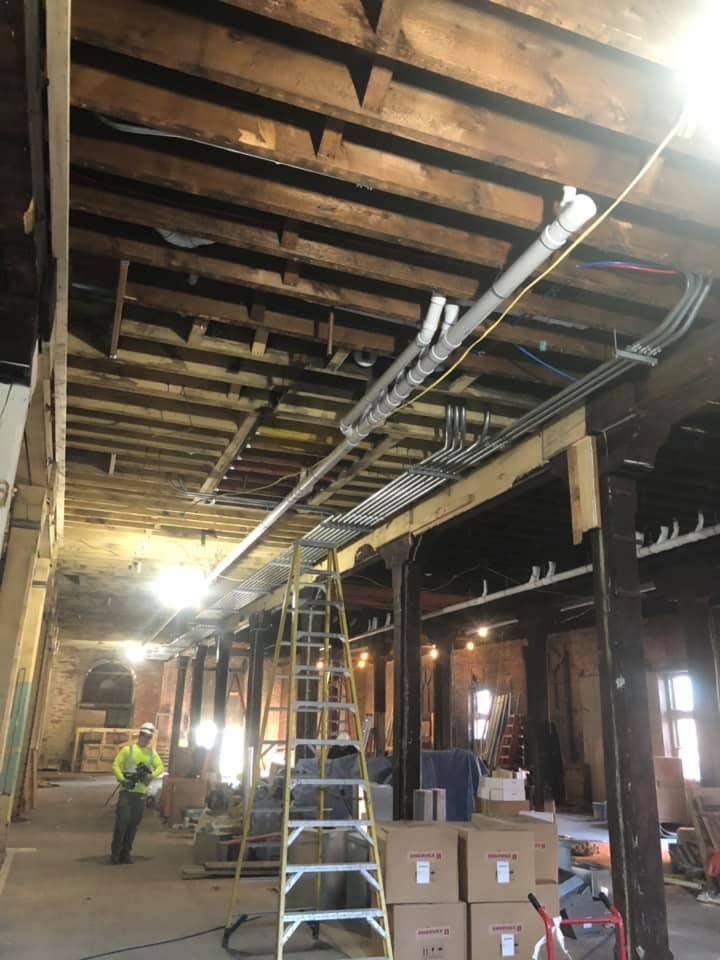 Worker on ladder in unfinished building with exposed beams and stacked boxes.